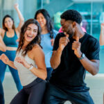 A large group of adults are seen in a studio together, as they participate in a Zumba fitness class.  They are each dressed comfortably and appear to be having fun as they dance and keep their heart rates up.